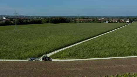 Aerial-view-at-a-tractor-driving-on-a-country-road-next-to-a-growinggreen-field-with-a-village-in-the-background