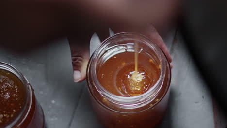 Close-up-View-of-Hand-Holding-a-Glass-Jar,-Pour-and-Fill-it-with-Golden-Fresh-Liquid-Extracted-Bee-Honey,-Traditional-Apiculture-Production-and-Beekeeping-in-Bee-Farm