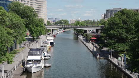 Aufnahme-Des-Rideau-Kanals-Neben-Dem-Gebäude-Des-Kanadischen-Senats-An-Einem-Sonnigen-Sommertag-Mit-Mehreren-Booten-Im-Bild---4k