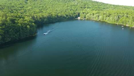 Aerial-drone-shot-flying-high-over-a-motor-boat-pulling-a-water-skier-along-the-lake-shore-on-a-sunny-day-in-New-Hampshire,-USA