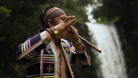 orbit-shot-of-Indigenous-person-playing-Kubing-tribal-instrument,-behind-is-a-waterfall-while-standing