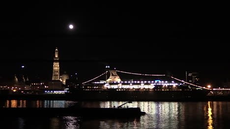 a-cargo-boat-passing-slowly-under-a-full-moon-at-the-cityscape-of-Antwerp