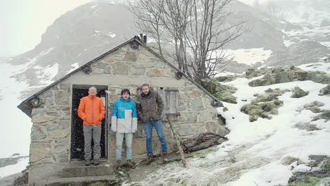 Three-hikers-pose-for-a-selfie-timer-photo-in-front-of-an-old-stone-cabin-in-the-Pyrenees-mountains-in-a-snow-storm-blizzard-in-daytime