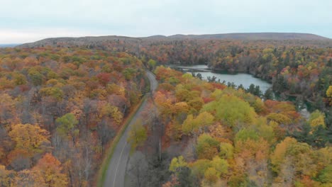 Slow-aerial-footage-to-the-left-over-a-car-on-a-curvy-road-in-an-autumn-forest-beside-Pink-Lake-in-Gatineau-Quebec-
