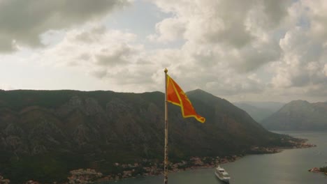 Static-shot,-of-a-flag-waving,-in-front-of-mountains-and-a-large-ferry-on-a-sea,-on-a-cloudy-day,-in-Kotor,-Montenegro