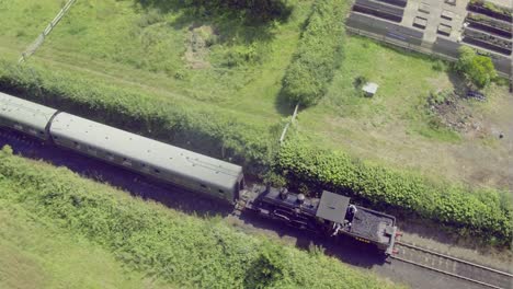 Overhead-aerial-view-of-a-steam-locomotive-pulling-carriages-over-a-level-crossing-into-Bodiam-train-station,-East-Sussex,-England