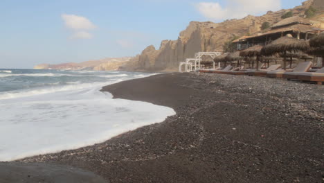 Tracking-shot-of-a-black-sand-beach-with-a-beach-resort-in-the-foreground-and-white-volcanic-cliff-formations-in-the-background