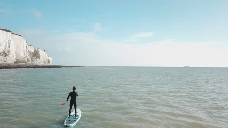 Junger-Mann-Steht-Beim-Stand-Up-Paddle-Boarding-Im-Meer-Mit-Weißen-Klippen-Von-Dover-Und-Blauem-Himmel-Im-Hintergrund