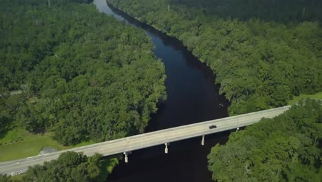 Drone-shot-over-a-bridge-on-a-river,-with-a-black-pickup-driving-over-it-on-a-sunny-day