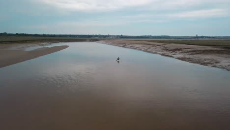 Man-kayaking-on-meandering-river,-drone-pans-up-to-reveal-beautiful-landscape-in-the-background