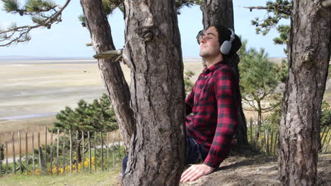 Shot-of-an-authentic-hipster-man-listening-to-music-with-headphones-whilst-sitting-in-the-sun-against-a-tree-trunk