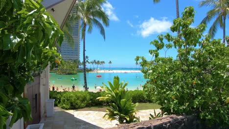 View-From-Resort-Wall-And-Bushes-Showing-Pool-Surrounded-By-Sand-With-Hotel-In-The-Background,-Waikiki,-Hawaii