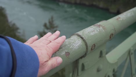 Person's-hand-running-down-railing-of-bridge-with-green-fast-flowing-river-below