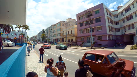 Footage-of-Havana,-Cuba-on-a-sunny-and-windy-day-with-people-waiting-and-walking-on-the-sidewalk-with-usual-traffic-driving-up-and-down-the-avenue