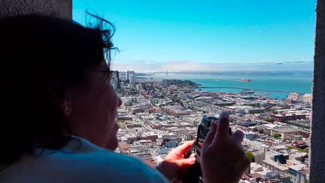Woman-taking-a-picture-of-SF-bay-from-top-of-Coit-Tower-looking-over-San-Francisco,-California,-USA