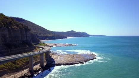 Aerial-footage-of-a-stunning-long-curvy-Bridge-next-to-the-sea-hightlights-the-Grand-Pacific-Drive-with-south-pacific-ocean-and-blue-sky-view-on-a-bright-sunny-day,-Sydney,-Australia