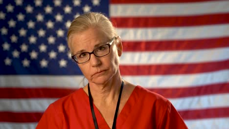 Medium-tight-portrait-of-nurse-looking-concerned-and-sad-putting-her-glasses-on-with-American-flag-behind-her