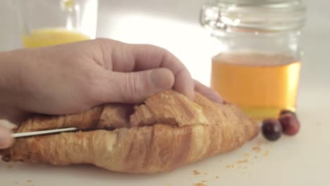 Hand-cutting-open-fresh-baked-croissant-with-honey-and-orange-juice-background-close-up-shot