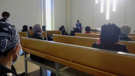 Hombre-Africano-Predicando-En-Un-Funeral-Sombrío-Mientras-La-Congregación-Está-Sentada-En-Bancos,-Rezando,-Escuchando-Y-Presentando-Sus-Respetos