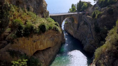 Fiordo-di-Furore-arch-bridge-with-a-boat-entering-the-passageway-to-village,-Amalfi-coast-in-Salerno-Italy,-Aerial-approach-down-shot