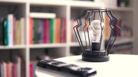 A-stylish-lamp-stands-on-a-white-table,-a-black-remote-control-is-next-to-it,-in-the-background-there-are-books-as-a-blurred-background