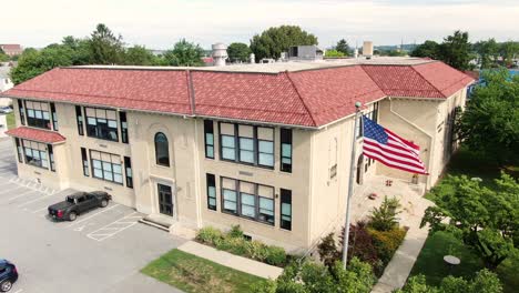 Exterior-of-American-public-school-building,-USA-flag-flying-in-wind,-establishing-shot,-aerial,-Spanish-style-architecture-with-terracotta-roof