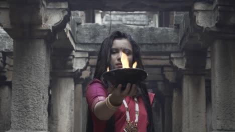 beautiful-Indian-girl-in-old-stepwell-wearing-traditional-Indian-red-saree,-gold-jewellery-and-bangles-holding-fire-plate