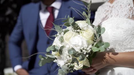 Bride-with-beautiful-flower-bouquet-in-hand-and-amazing-white-dress,-man-in-blue-suit-standing-beside-her-out-of-focus