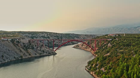 wide-aerial-shot-of-a-big-red-bridge-on-a-beautiful-mountain-scenery-on-sunset,-camera-moving-forward
