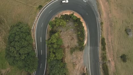 Aerial-birds-eye-drone-view-of-a-white-car-driving-round-a-bendy-road-in-the-hills-of-new-zealand