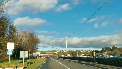 Driving-Kinsale-pier-road-on-a-sunny-day,-passing-docked-fishing-boats-and-people-walking