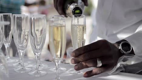 Close-up-shot-of-a-black-man-pouring-champagne-sparkling-wine-in-tall-transparent-glasses-at-a-wedding-with-a-blurred-background,-social-concept