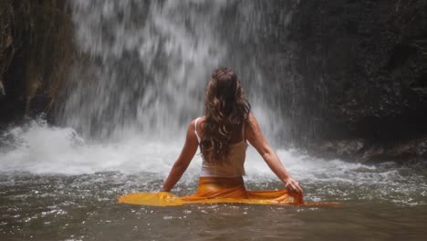 A-slow-motion-shot-of-a-young,-attractive-woman-dancing-in-the-water-with-a-yellow-skirt-with-a-small-waterfall-behind-her