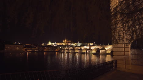 The-Prague-Castle-and-Charles-bridge-over-river-Vltava-in-the-historical-centre-of-Prague,-Czechia,-lit-by-lights-at-night,-shot-from-the-other-side-of-the-river,-zooming-through-willow-branches
