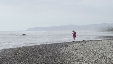 Tourist,-Der-An-Einem-Nebligen-Morgen-Am-Strand-Von-Olon,-Ecuador,-Am-Ufer-Feiner-Steine-Mit-Tosenden-Wellen-Steht