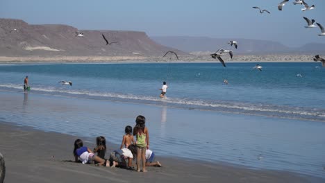 Establishing-shot,-Children-playing-on-the-beach-of-San-Juanico,-California-Sure,-Mexico,-a-kid-a-with-selfie-stick-chasing-the-birds-in-the-background