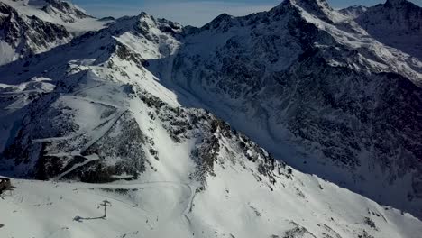 Establishing-shot-of-Tourist-Skiers-riding-down-ski-track-rocky-mountain-slope-in-Val-Thorens,-French-Alps---Aerial-High-Fly-over-descending-shot