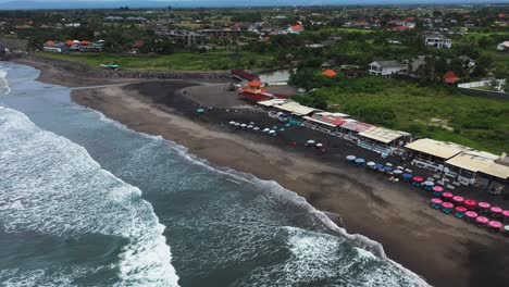 Pererenan-Beach-in-Bali-Indonesia-with-beach-bars-and-cafes-by-the-ocean-waves,-Aerial-dolly-in-reveal-shot