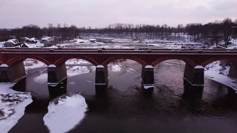 Aerial-view-of-old-red-brick-bridge-across-the-Venta-river-in-Kuldiga,-Latvia-in-overcast-winter-day,-Venta-rapids-in-background,-wide-angle-drone-shot-moving-backwards