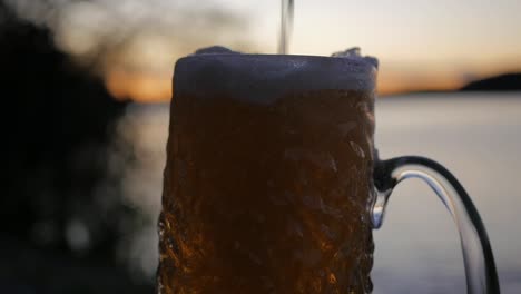 Während-Die-Sonne-über-Dem-Schwedischen-Archipel-Untergeht,-Wird-In-Zeitlupe-Ein-Kaltes-Lagerbier-In-Ein-Geriffeltes-Bierglas-Eingefüllt