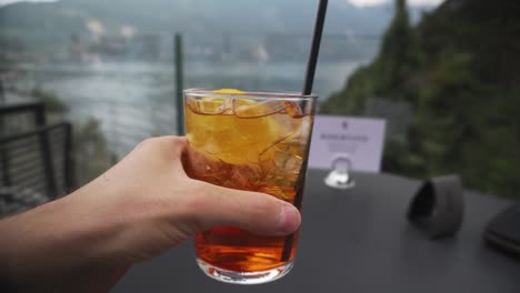 Man-rising-glass-of-aperol-spritz-from-table-in-restaurant-by-Garda-lake,-Italy