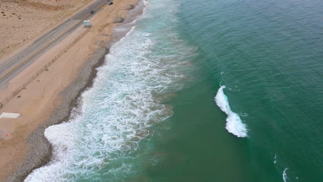 Flying-Along-Shoreline-in-Malibu,-Aerial-Shot-Ocean-Waves-Crashing-against-Sand,-Cars-Moving-along-Highway