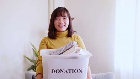 Porträt-Einer-Jungen-Asiatischen-Frau,-Die-In-Die-Kamera-Schaut-Und-Eine-Box-Mit-Kleidung-Für-Spenden-Hält