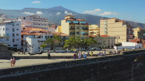 The-seaside-skyline-of-Puerto-de-la-Cruz,-Canary-Islands,-Spain,-tourists-walking-on-its-stone-harbor-walkway-enjoying-the-view-on-a-sunny-day,-Pico-de-Teide-mountain-in-the-background,-aerial-4K