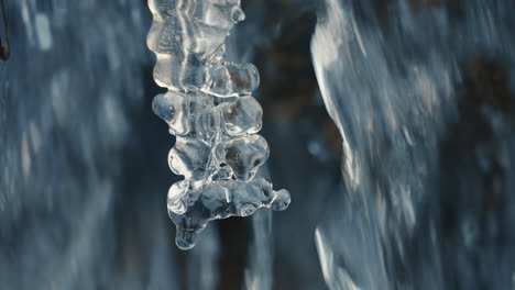 drop-falling-on-icicle-in-a-frozen-river-with-flowing-water-in-background,-close-up,-slow-motion