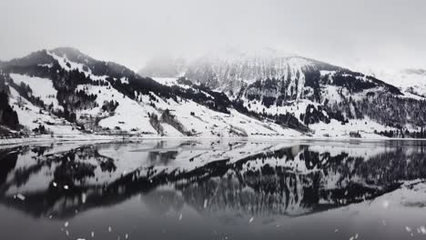 Snowy-mountain-landscape-reflects-in-clear-lake-water-during-snowfall,-aerial-drone-view
