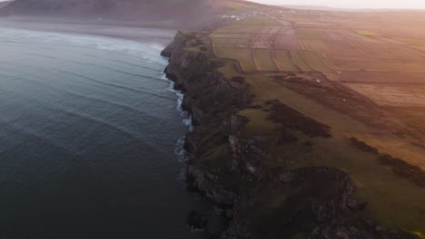 AERIAL:-Dolly-along-cliff-face-coastline-during-hazy-sunrise,-Rhossili-Gower,-4k-Drone