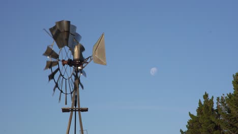 Old-windmill-turning-with-the-wind,-with-the-moon-in-a-blue-sky,-in-Patagonia,-Argentina