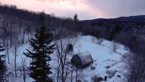 Drone-orbits-around-barn-in-snowy-mountains-of-Vermont-during-radiant-sunset