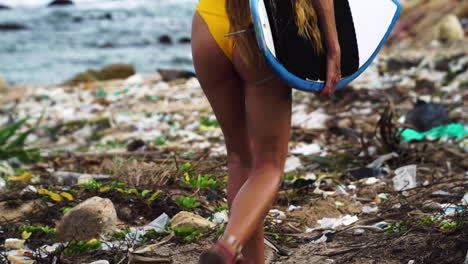 Woman-in-yellow-bikini-and-surfboard-walking-along-polluted-beach